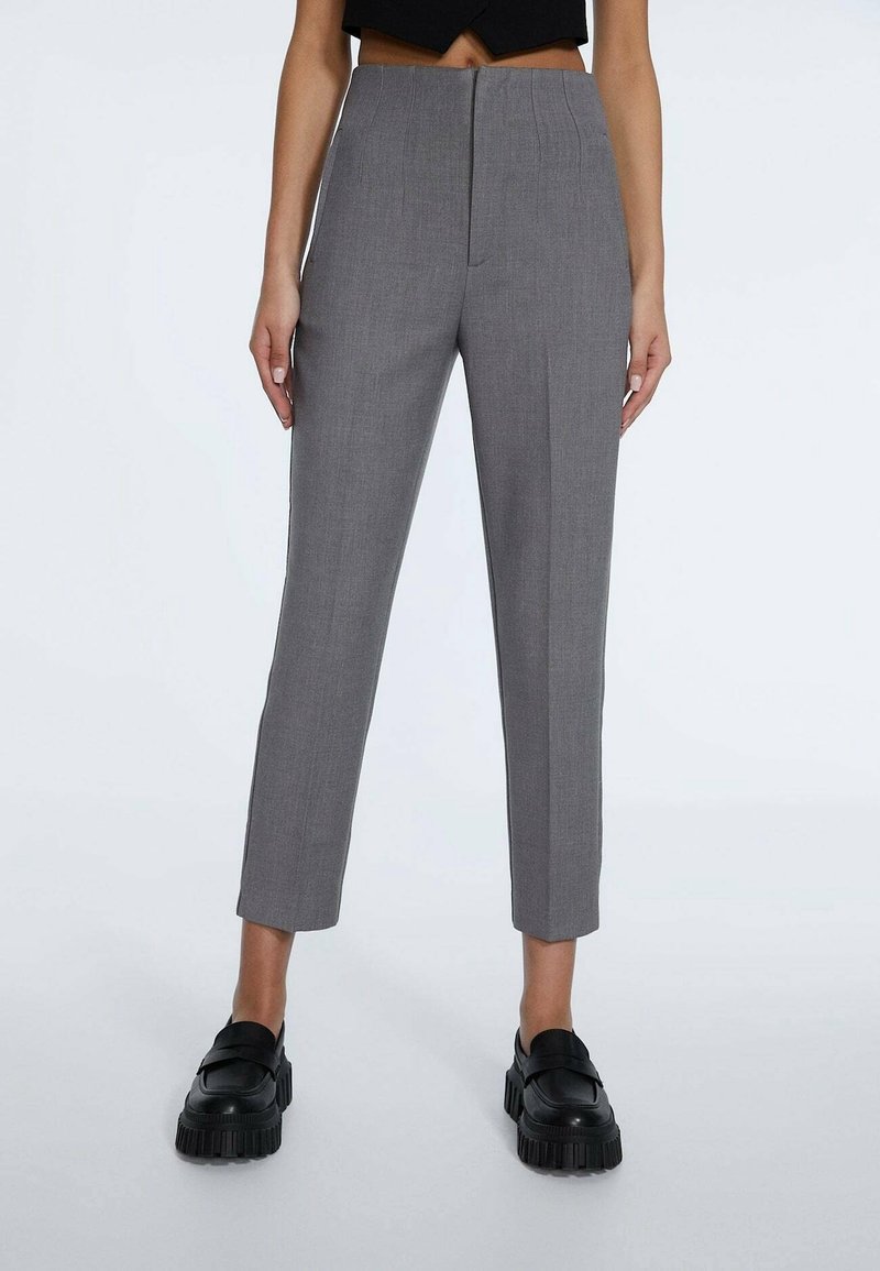 Femme portant un pantalon gris sur mesure taille haute, longueur cheville, et des mocassins noirs épais, debout devant un fond clair uni.