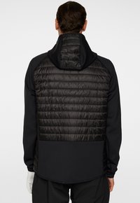 Svart jacka med en vadderad, vadderad rygg och huva. Släta, strukturerade ärmar och nederdel. Minimalistisk design, inga synliga logotyper eller detaljer.