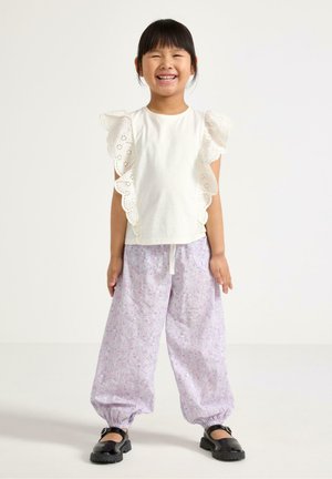 Smilende pige iført hvid rynket ærmeløs top, løstsiddende lyse lilla blomstrede bukser med elastiske manchetter og sorte sko mod ensfarvet baggrund.