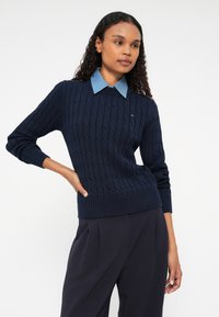 Tamsiai mėlynas megztinis su pynėmis, su raukšlėtu apykakliu ir rankogaliais, uždėtas ant šviesiai mėlynos marškinėlių su apykakle. Ant krūtinės yra nedidelis logotipas.