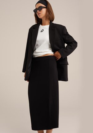 Zwarte oversized blazer, witte strak zittende t-shirt met logo, en zwarte midi rok. Soepele texturen, ontspannen pasvorm en minimalistisch ontwerp.