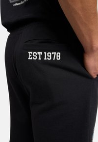 Black sweatpants featuring a back pocket with white embroidered text "EST 1978," smooth fabric, and a tapered shape.