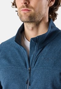 Blå zip-up jacka gjord av texturerad tyg. Har en hög krage, kontrasterande paneler och en broderad logotyp på bröstet.