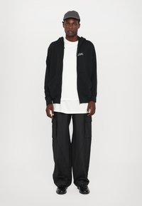 Black zip-up hoodie with front pockets, white T-shirt, black cargo pants, and black shoes. Gray cap completes the outfit.