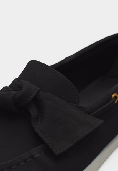 Μαύρο παπούτσι χωρίς κορδόνια με μεγάλο υφασμάτινο φιόγκο στην κορυφή, ραμμένες λεπτομέρειες και λευκή σόλα πάνω σε απλό φόντο.