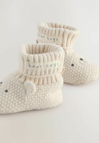 Caldi scaldamuscoli per neonati in crema lavorati a maglia, con polsini a coste, design a faccia d'orso e etichetta "abbracci d'orso". Caratterizzati da una morbida texture di lana e forma arrotondata.