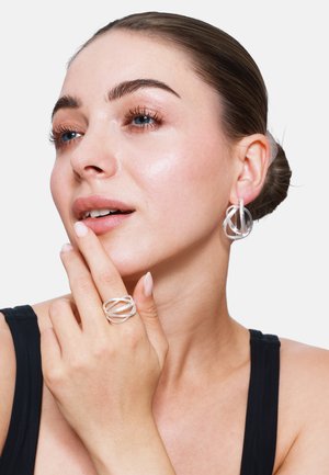 Zilveren sieradenset met verstrengelde oorbellen in de vorm van een ring met een spiraalontwerp. Model benadrukt de gestructureerde oppervlakken tegen een zwarte top.