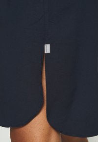 Robe navy avec un ourlet arrondi et une fente sur le côté. Fabriquée en tissu lisse, elle présente une étiquette avec le nom de la marque près de l'ourlet.