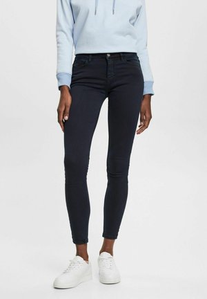Person iført mørke skinny jeans, lyseblå sweatshirt og hvide sneakers mod en ensfarvet hvid baggrund.