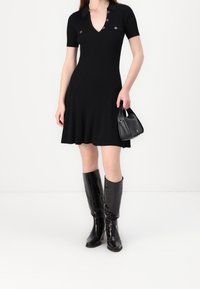 Robe en maille côtelée noire avec manches courtes et col en V, agrémentée de boutons-pression, associée à des bottes noires brillantes montantes jusqu'aux genoux et à un sac à main.