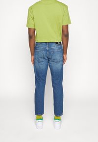 Grönt bomullströja, blå jeans med rak passform och vita sneakers. Framhävd gröna och gula strumpor synliga ovanför skosträngen.