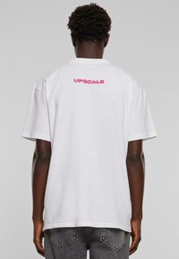 Hvid bomulds t-shirt med korte ærmer, med et pink "UPSCALE" grafisk motiv på bagsiden. Løs pasform og blød tekstur.