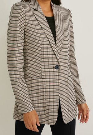 Femme portant un blazer pied-de-poule beige et noir avec un bouton noir unique, par-dessus un haut noir et un pantalon noir, debout devant un fond uni.