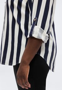 Χέρι και πλευρικό μέρος κορμού ενός ατόμου που φοράει πουκάμισο με κάθετες ναυτικές και λευκές ρίγες με τα μανίκια ανασηκωμένα και μαύρο παντελόνι.