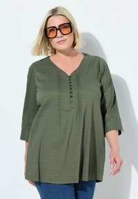 Olivgrüne Bluse aus strukturiertem Stoff, mit V-Ausschnitt, Knopfdetails und Dreiviertelärmeln. Kombiniert mit blauer Jeans.