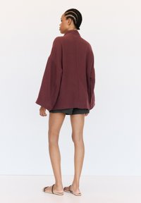 Veste oversized bordeaux en tissu texturé avec des manches larges, associée à un short noir et des sandales. La veste présente un col montant et une coupe ample.