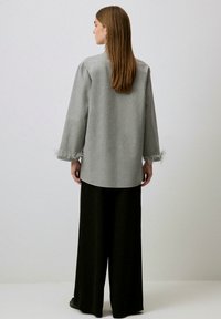 Palton gri, tailored, cu mâneci lungi, accentuat cu pene la manșetă, asortat cu pantaloni negri largi. Material neted și structurat.