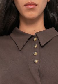 Polo marron en tissu texturé, avec un col pointu et quatre grands boutons marron clair le long de la patte de boutonnage.