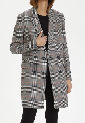 Femme portant un long manteau noir et blanc à motif pied-de-poule avec des lignes rouges, à double boutonnage, par-dessus un haut noir et un pantalon noir.