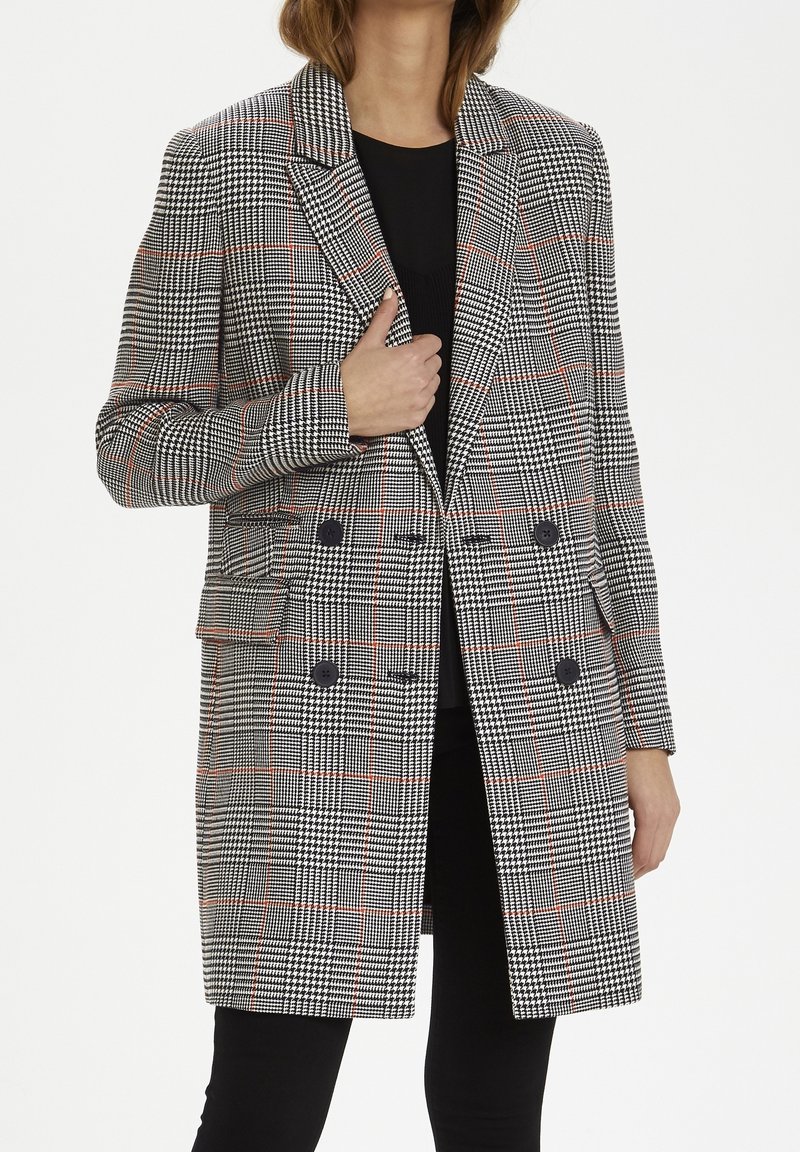Femme portant un long manteau noir et blanc à motif pied-de-poule avec des lignes rouges, à double boutonnage, par-dessus un haut noir et un pantalon noir.