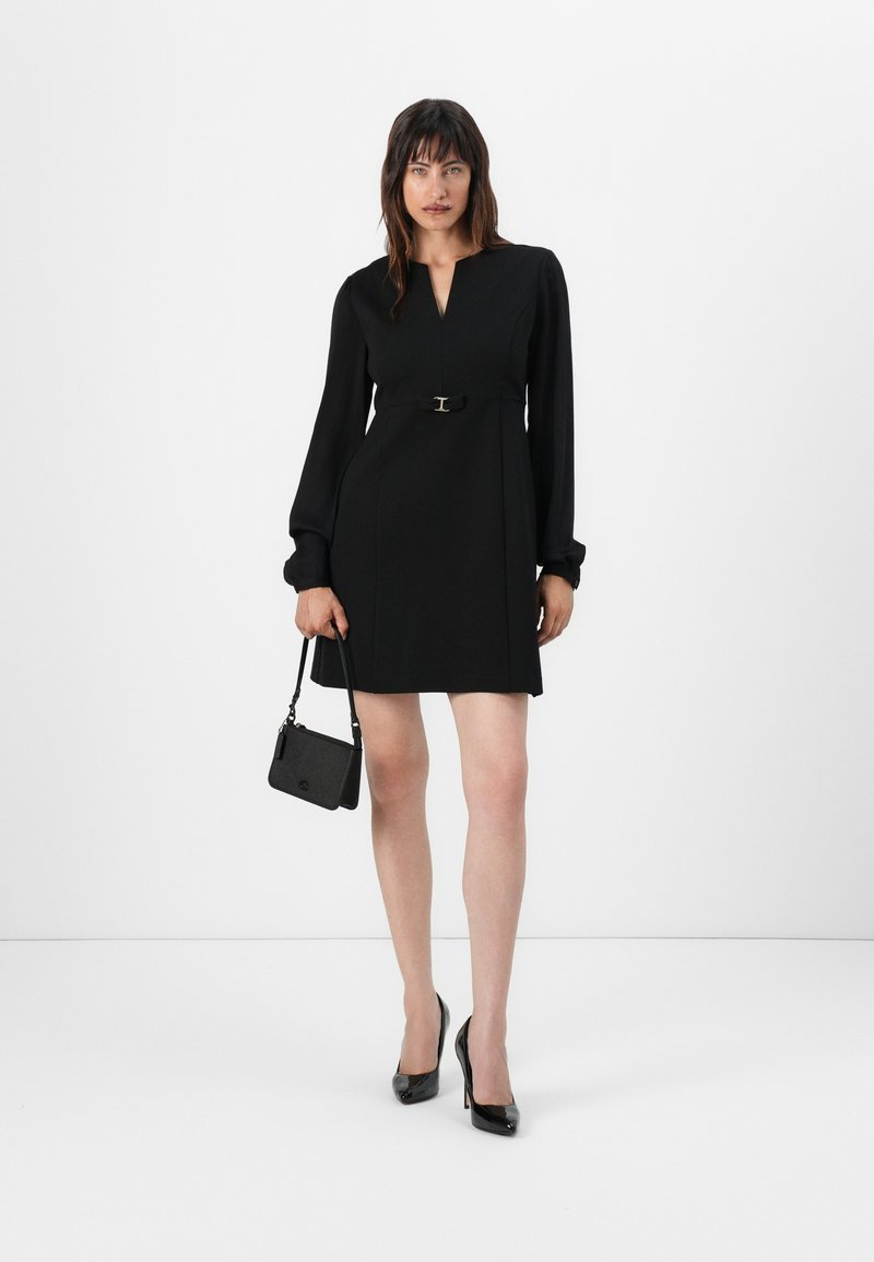 Abito nero con maniche lunghe, scollatura a V, vita aderente con una fibbia argentata in vita. Abbinato a tacchi alti neri e una piccola borsa a tracolla.