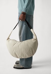 Bolso de hombro acolchado beige con un diseño curvado, asas negras y un cierre superior. Lleva puesto una persona con un suéter azul claro y jeans azules.
