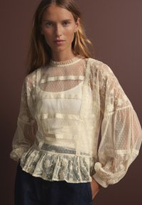 Blusa plissettata avorio con dettagli in pizzo a pois e floreale. Caratterizzata da maniche lunghe trasparenti, tessuto testurizzato e orlo arricciato.