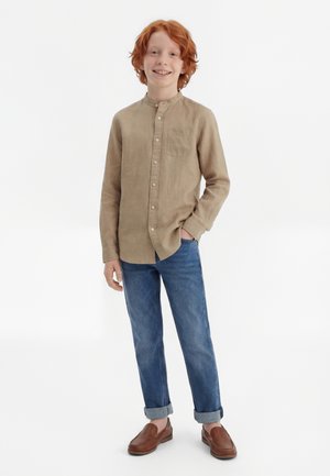 Smiling red-haired boy stands with one hand in pocket wearing beige button-up shirt, rolled-up blue jeans, and brown loafers against white background.