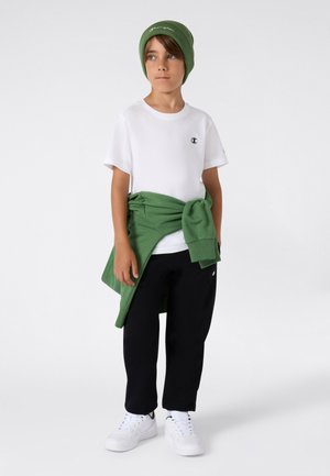 White short-sleeve t-shirt with logo, green sweatshirt tied at waist, black pants, and white sneakers. Simple, casual design.