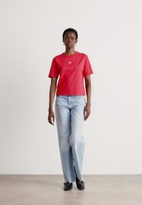 Rød kortærmet t-shirt med et lille logo, lyseblå jeans og sorte sko. Stofet virker glat, og pasformen er afslappet.