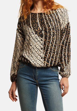 Vrouw draagt een blouse met abstract patroon in zwart, beige en bruin, lange mouwen en knopen, gecombineerd met een blauw spijkerbroek, staand met ontspannen handen.