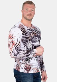 Witte lange mouw shirt met zwarte vleugel- en kroonafbeelding, met spetters van rood en bruin. Gepaard met blauwe jeans staat het model met een neutrale uitdrukking.