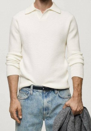 Mężczyzna w białym swetrze z kołnierzem i jasnoniebieskich dżinsach, trzymający w prawej ręce szarą tkaninę.