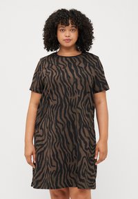 Curly-haired woman wearing a short, brown dress with a black zebra stripe pattern, standing against a plain white background.