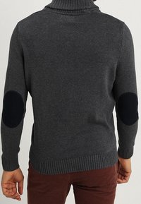 Uomo che indossa un maglione grigio scuro a collo alto con toppe nere sui gomiti e pantaloni marroni, visto da dietro.