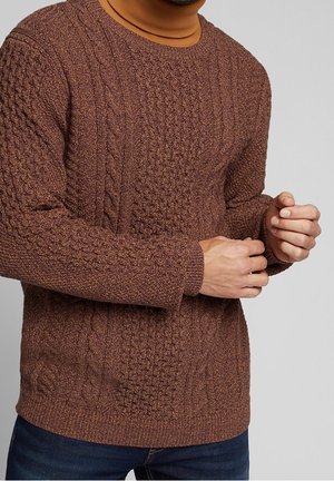 Brauner Kabelstrick-Pullover mit rundem Ausschnitt, strukturiertem Stoff und gerippten Bündchen. Kombiniert mit einem hellbraunen Rollkragenpullover darunter.