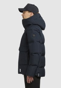 Marineblå puffjakke med en stor hætte, lynlås foran og to frontlommer. Har en struktureret stofkvalitet og et mærket patch på ærmet.