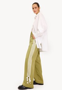 Wijdvallende, olijfgroene broek met witte strepen, gecombineerd met een lange, oversized witte blouse. Zwarte schoenen met een gestructureerde zool.