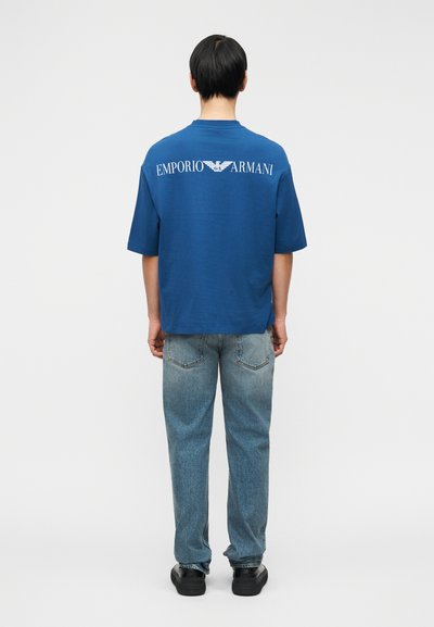 Μπλε βαμβακερό μπλουζάκι με κοντά μανίκια, με λευκό λογότυπο "Emporio Armani" στην πλάτη. Συνδυάζεται με ανοιχτό μπλε τζιν παντελόνι.