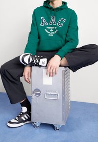 Grön huoodad sweatshirt med vit text, svarta cargobyxor och svarta och vita sneakers. Sitter på en silverfärgad metallåda med hjul.