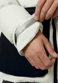 Tessuto bianco della giacca al polsino con una striscia riflettente argentea; materiale scuro nel corpo della giacca; si vede una mano che aggiusta il polsino.