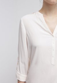 Blouse beige clair avec un col en V, manches longues retroussées et fermeture à boutons. Tissu lisse avec un subtil éclat.
