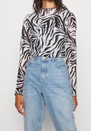 Person iført en sort-hvid zebra mønstret langærmet top med transparente ærmer, der er stoppet ned i højtaljede lyseblå denimjeans.