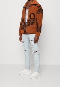 Brun, fluffig zip-jacka med svart logodesign, parat med slitna ljusblå jeans och vita sneakers.