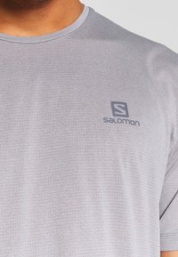 Grå sporttröja gjord av andningsbart tyg. Har rund halsringning, korta ärmar och en liten logotyp på övre vänstra bröstet.