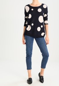 Navy knitted sweater with large cream circles, round neckline, and long sleeves, paired with slim-fit denim jeans and black flat shoes.
