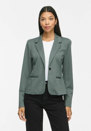 Femme aux longs cheveux foncés portant un blazer vert sauge cintré sur un haut blanc et un pantalon noir, debout devant un fond blanc uni.