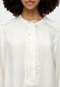 Weiße Bluse mit einem strukturierten Fransenbesatz entlang der Vorderseite und des Kragens, mit einer gebundenen Schlüsselöffnung. Weicher Stoff mit glattem Finish.