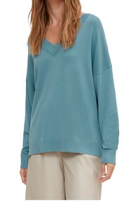 Femme portant un pull ample à col en V de couleur turquoise et un pantalon beige, visible des épaules jusqu'à mi-cuisse, debout avec les bras détendus.