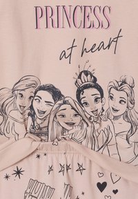 Light pink cotton garment featuring black line drawings of five princesses, with "PRINCESS at heart" text and star patterns below.